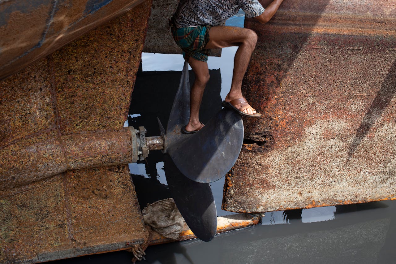 Man in sandals standing on a large rusty ship propeller outdoors during the day.
